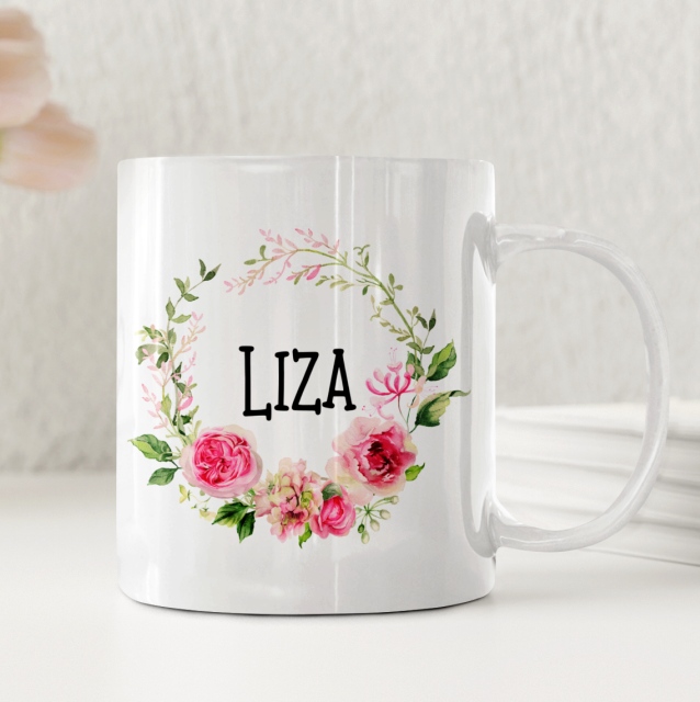 Vit keramikmugg med en rosa blomsterkrans och ett personligt namn i mitten.