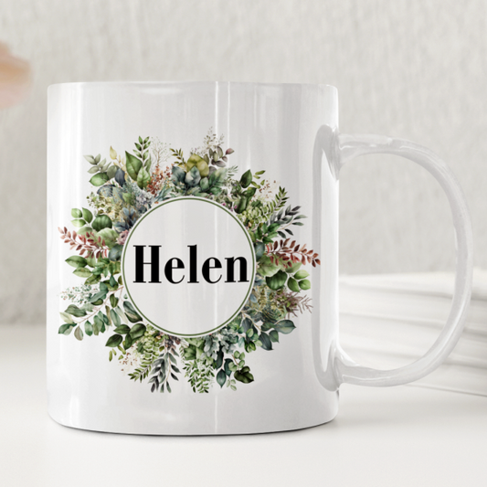 Vit keramikmugg med en cirkel av gröna blad och ett personligt namn i mitten.